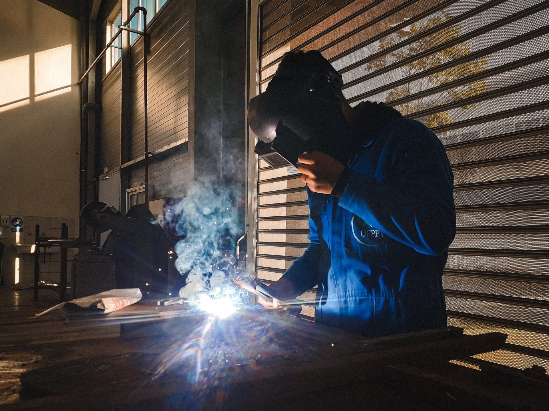 A welder in blue overalls at work, sparks and smoke rising from the joint
