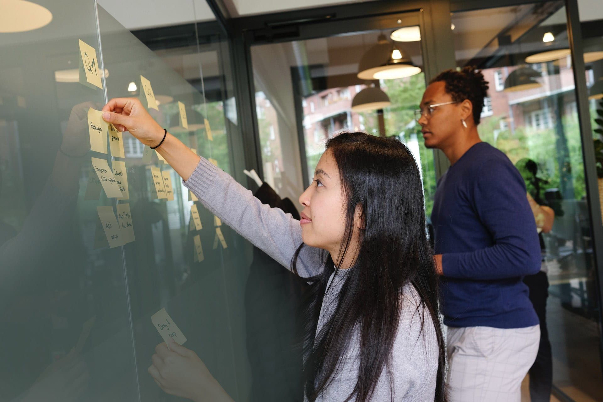 A woman sticking notes on a glass wall while a colleague looks on
